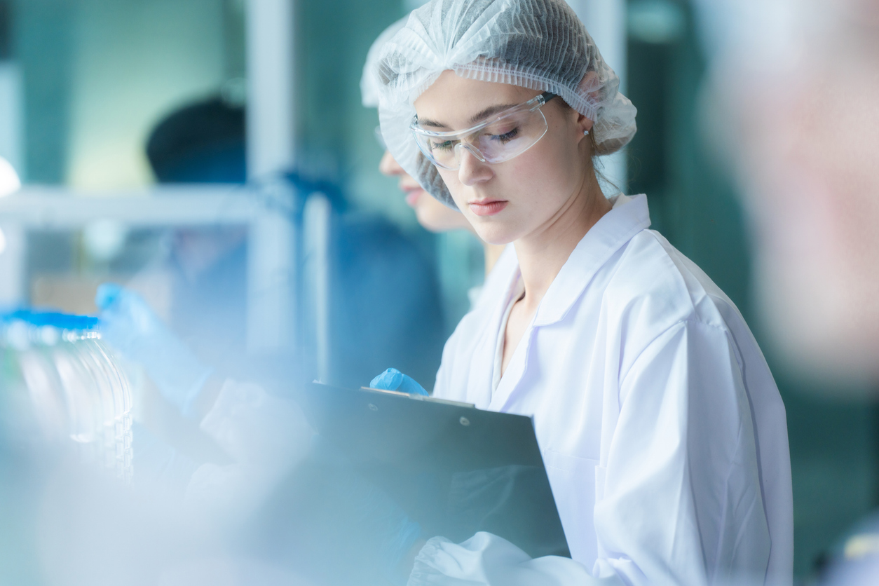scientist worker checking the quality of water bottles on the machine conveyor line at the industrial factory. Female worker recording data at the beverages manufacturing line production.